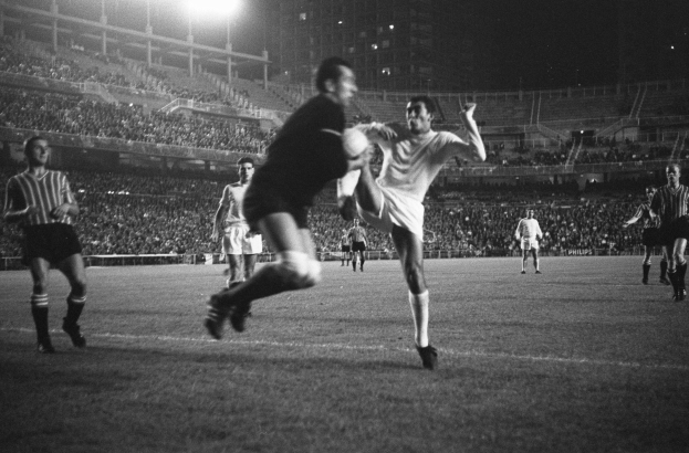 Schwarzes Foto von Männern, die Fußball in einem beleuchteten Stadion spielen, umgeben von Gebäuden.