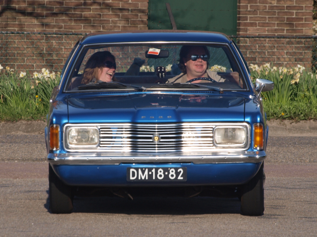 Ein Mann und eine Frau fahren in einem blauen Auto öffentlich, beide tragen Sonnenbrillen und löcheln, mit einer Backsteinwand, einer grünen Tür, blühenden Pflanzen und Maschendraht im Hintergrund.