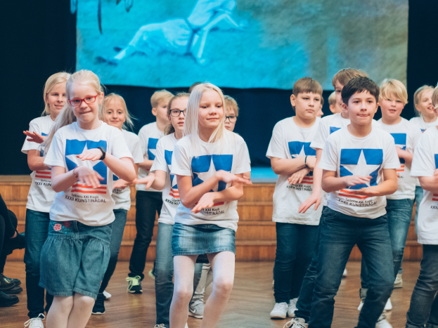 Eine Gruppe von Kindern in weißen Hemden und blauen Shorts, einige mit Brille, tanzt auf einer Bühne mit Zuschauern auf der linken Seite und einem Bildschirm im Hintergrund, bei einem Tanzwettbewerb.