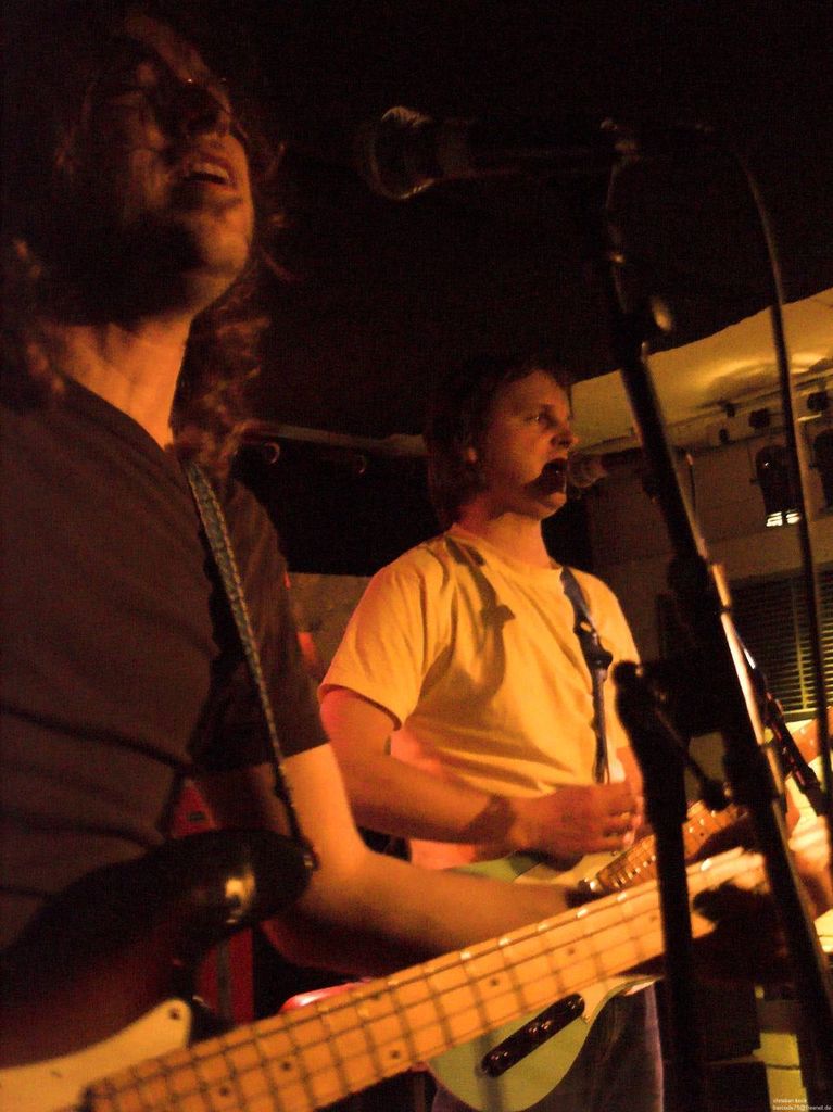 Eine Person in einem weißen Hemd singt in ein Mikrofon, während sie eine grüne Gitarre spielt und neben einer anderen Person in einem schwarzen Hemd und Brille steht, die ebenfalls singt und eine schwarze Gitarre spielt, mit Scheinwerfern und anderen Instrumenten im Hintergrund.