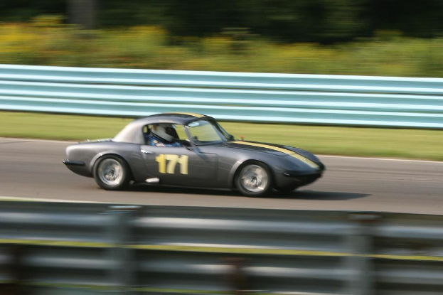 Ein Auto mit einem Helm tragenden Fahrer bewegt sich auf einer Straße, die von Barrieren gesäumt ist, mit Bäumen im Hintergrund.
