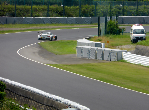 Ein Rennwagen fährt auf einer Rennstrecke mit einem Polizeiwagen im Hintergrund, umgeben von Gras, Pflanzen, Bäumen und einem Zaun, mit einem Fahrzeug auf der rechten Seite und Text unten.