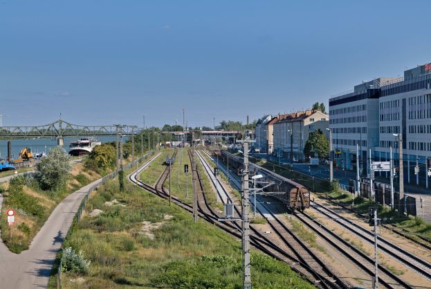 Ein Zug fährt auf Schienen neben einer Stadtlandschaft mit Infrastruktur, Gebäuden, Grünflächen, Straßen, einer Brücke, Wasser und einem bewölkten Himmel.