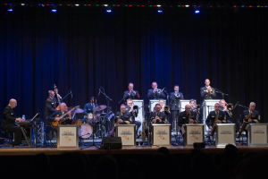 Eine Gruppe von Musikern auf der Bühne, die Instrumente spielen, mit Mikrofonen und Ständern, Schilder mit der Aufschrift "The New Orleans Jazz Band", ein Vorhang im Hintergrund und ein Publikum darunter unter Bühnenlichtern.