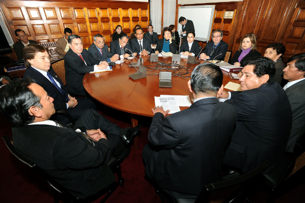 Rundtischgespräch mit Delegierten, die über Mikrofone, Papiere, Kabel und Dateien auf dem Tisch sowie eine hölzerne Wand im Hintergrund diskutieren.