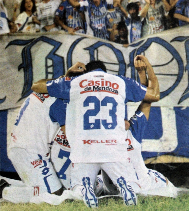 Gruppe von Fußballspielern, die auf dem Boden feiern, mit jubelnden Zuschauern im Hintergrund und einem Banner mit Text im Hintergrund.