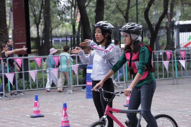 Zwei Frauen mit Helmen fahren auf einer von Verkehrskegeln gesäumten Straße Fahrräder, mit Menschen, Bäumen und einem Geländer im Hintergrund und einer Tafel mit Schrift auf der linken Seite.