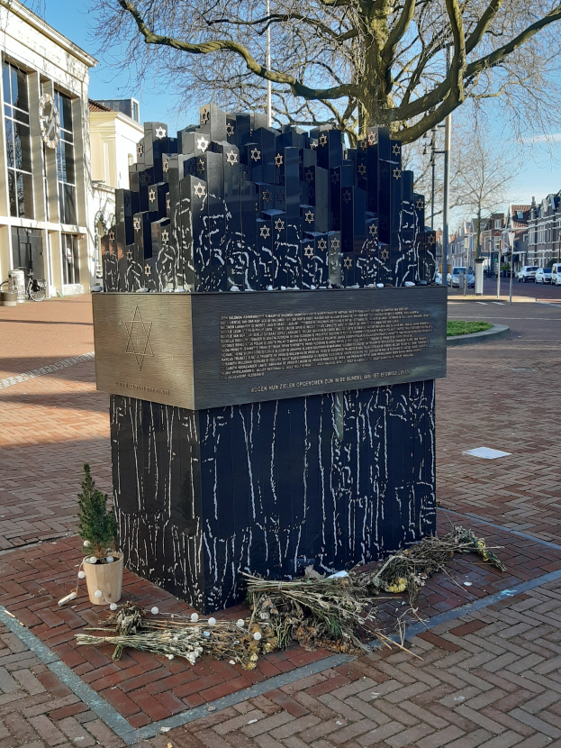 Denkmal für Holocaust-Opfer in einem Stadtplatz mit einem Baum, Topfpflanze, verstreuten Blättern und umliegenden städtischen Elementen.
