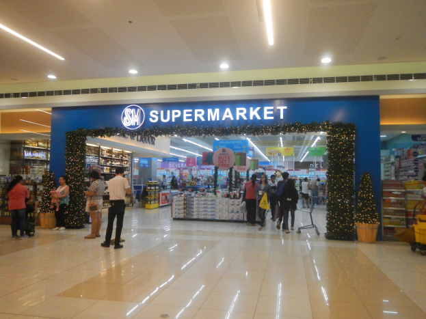 Gruppe von Menschen vor einem Supermarkt mit Einkaufstägern, einigen mit Einkaufstägern, mit einem Einkaufswagen, Topfpflanze, Regalen, Schildern und Deckenbeleuchtung.