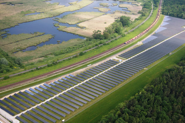 Luftaufnahme einer Solarpark mit Solarpanelen in einem Feld, umgeben von Bäumen, Gras, Pflanzen und Wasser, mit einem Zug auf einer nahen Bahnstrecke.