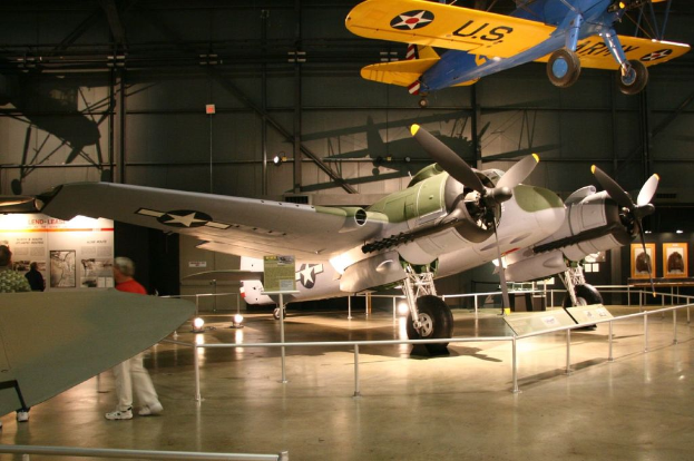 Ein Innenblick in einen Hangar mit Flugzeugen, eines hängt von der Decke, Lampen auf dem Boden, Menschen in der Nähe eines Zauns und eine Tafel mit Text an der Wand, beleuchtet von nahen Lampen.