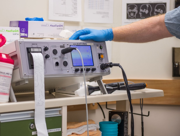 Eine Person in blauen Handschuhen arbeitet an einem medizinischen Gerät auf einem Tisch in einem unordentlichen Krankenhauszimmer mit Papieren an der Wand.