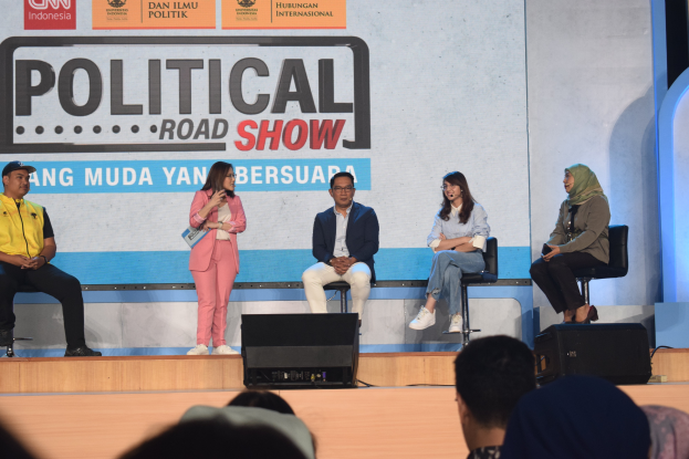 Eine Gruppe von Menschen auf einer Bühne sitzend, mit einer Frau in der Mitte, die ein Mikrofon hält, und einem Publikum im Vordergrund, mit einem Banner im Hintergrund, das 'Politische Straßen-show 2019' lautet.