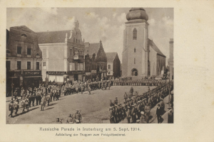 Ein Schwarz-Weiß-Foto eines Umzugs in Insterburg am 5. September 1914 mit vielen Menschen, Gebäuden und einem bewölkten Himmel sowie einem Text am unteren Rand.