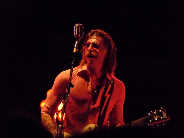 Ein Mann mit Brille steht singend da und hält eine Gitarre, mit offenem Mund.