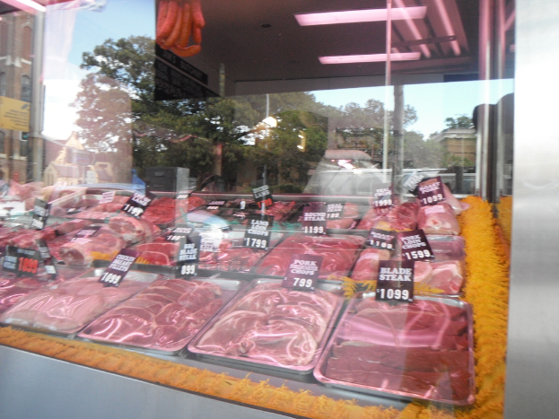 Eine Glasvitrine in einem Metzgerladen zeigt Tabletts mit Fleisch und Preisschildern, mit Gebäuden und Bäumen im Fenster und der Decke des Raums oben.